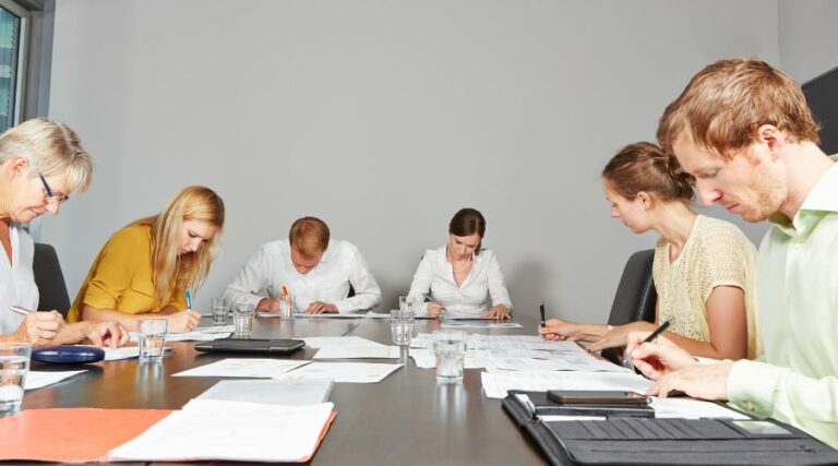L'immagine rappresenta un gruppo di persone al lavoro, impegnate in un assessment center. Ci sono uomini e donne seduti intorno al tavolo, che stanno riflettendo sulla prova che gli è stata assegnata.